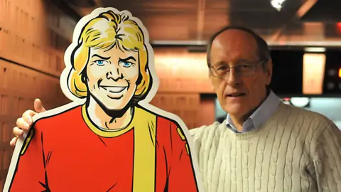 Barrie Tomlinson, standing next to a cardboard cut-out of Roy of the Rovers, with his arm round it. The cut-out shows a cartoon footballer in a red shirt, with a yellow stripe and long blonde hair and blue eyes. Barrie has on a white jumper, glasses and a blue shirt.