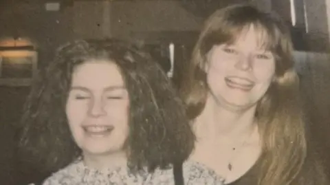 Diane Barnes An old photo of two young women on a night out. They are grinning at the camera and standing beside each other. One has dark hair and the other has brown hair. They are standing in a darkly lit indoor venue. 