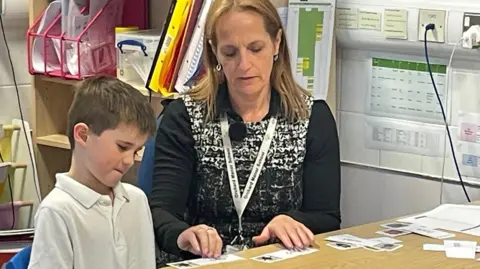 Primary school student Lucas chooses the correct words as a teacher tells them. They are both seated at a desk in a classroom.