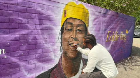 A man holding a pen colouring in the teeth of a mural on a wall. The wall is purple, and celebrates Eddie Kinuthia with a drawing of him