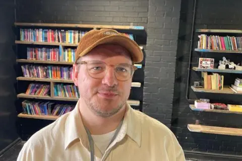 Sally Fairfax A man in glasses, a brown cap and a cream shirt is standing in front of bookshelves, full of slim, colourful volumes. They are lining an interior wall built of black-painted bricks. 