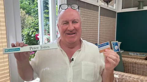 Kate Bradbrook/BBC Peter holds up his former desk ID in one hand and an old-fashioned blue and beige Barclaycard in the other. He smiles at the camera wearing a white linen short sleeve shirt.