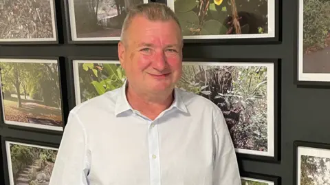 ELLIOTT GREEN/BBC Dean, also stood in the exhibition. In the photos behind him, we can see shots of trees, pathways, and stairs.