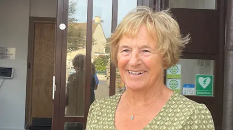 Joan Simon has short blonde hair and a pale green cap sleeved dress. She is standing outside the open front door of the school and is smiling at the camera.