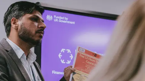 A man in a grey suit stood in a classroom at the front, speaking to the class