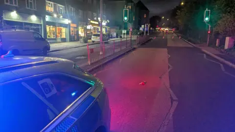 A police car is on the road on the left of the picture. The road has traffic lights in the middle and a line of cones to stop traffic. On the left are rows of shops. The scene is illuminated by a blue police light.