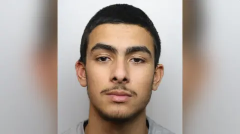 Tyler Wilkins with short dark hair and a slight beard wearing a grey top in a police custody photo