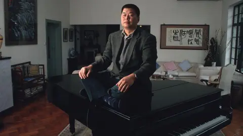 Getty Images Jimmy Lai, wearing a suit jacket and vest, sits above a grand piano at his home from a picture taken in 1993