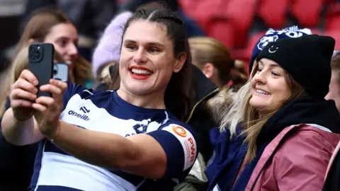 Ilona Maher poses for a selfie with a fan during her Bristol Bears debut . Ilona wears the blue and white striped Bristol Bears kit and red lipstick. She smiles as she holds a phone out for a photo with a young woman in a pink coat and black beanie. 