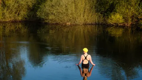 A female wild swimmer stands in a lake by herself wearing a black swimming costume. She wears a yellow swimming cap and has her back to the camera.