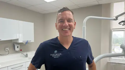 BBC Dr Mike Hesketh, wearing navy blue dental gown in a surgery, smiling