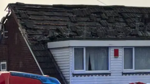 The burnt roof of a house, with many missing and loose roof tiles. The top of a fire engine can be seen in the bottom of the picture