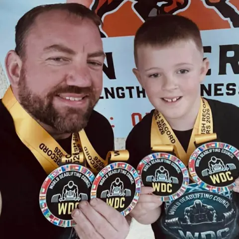 Stuart Whyte Stuart Whyte has a beard and short dark hair. He is wearing a black T-shirt. His son Konnan, who is next to him, has short dark hair. They are smiling broadly and displaying their medals.