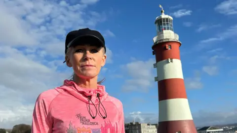 Nicky Putman is wearing a pink running jumper and a black cap on a bright sunny day in Plymouth. She is stood on grass looking straight at the camera. There is a lighthouse and the sea in the background.