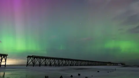 BBC Weather Watchers/lee A long pier cast in shadow because of the sky. Above, streaks of green and pink light illuminate the sky. The sea surrounding the pier is calm.