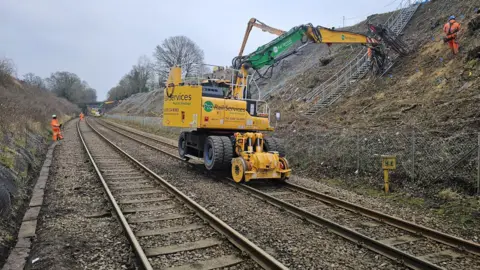 A yellow excavator moves down the track repairing the embankment between Guildford and Gatwick. Workers in hi-vis stand in the background.