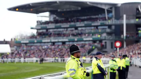 PA Media Police in yellow hi-vis jackets stand next to a railing by the green racetrack, with stands packed with thousands of spectators around them.