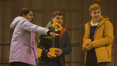 Children’s Theatre Partnership/Rose Theatre Three actors on stage holding sunflowers. A girl in a purple coat points to the right while two boys smile watching her