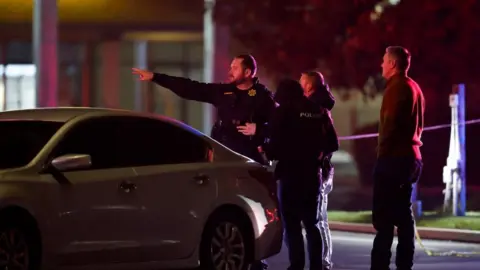 Police officers - one of them pointing with the outstretched arm, stand by a car near the scene in Stockton 