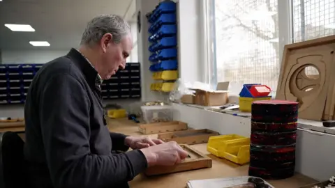 Neill Hunter working at a bench in a small workshop, handling wooden pieces, with storage trays and tools arranged along the wall and by a window. 