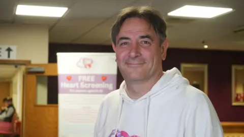 Jamie Niblock/BBC Karl Lansley, wearing a light grey hoodie with a pink logo on the chest for the Carli Lansley Foundation. Karl is stood in front of a white pull-up banner for the foundation. Karl has dark brown hair with a centre parting, and he is smiling at the camera.