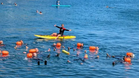 Students learn to float in the water while an instructor sits on a yellow surfboard. The instructor is wearing a wetsuit and has their arms outstretched.