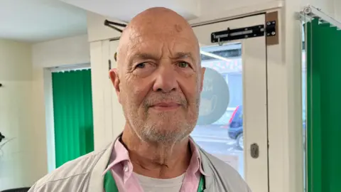 Malcolm looking into the camera and smiling slightly. He's wearing a grey jacket with a pink collared shirt underneath. He's stood in the doorway to the foodbank, with the window just showing a bit of the street beyond.