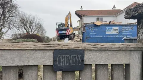 John Fairhall/BBC A digger in the distance on top of a mound as it demolishes a house. The house is white with a red roof. The entrance gate into the house is at the bottom of the picture with the name "Cheneys" on a black slate on the fence.