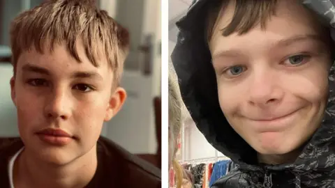 Max Dixon and Mason Rist. Mason is wearing a black crew neck jumper and has dark eyes and brown hair in a downward fringe. Max is wearing a black raincoat with his hood up and is smiling downwards, with pieces of his fringe peeking out. 