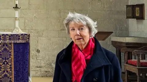 BBC Image of a woman wearing a red scarf and black coat, who is standing in front of an altar in a church.