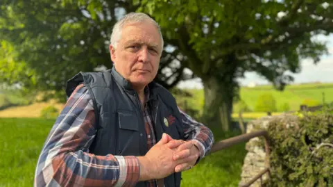 William Irvine, a man with short, grey hair, is leaning on a metal farm gate on a sunny day.  There are large, green trees and open fields behind him.   He is wearing a red, white and blue checked shirt under a navy bodywarmer.