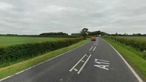 Google An A road in the middle of green fields. There is a red van driving away from the camera in the distance