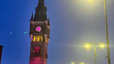 Foster with the North East Darlington's clock tower illuminated pink. Street lamps are lit up in the background.