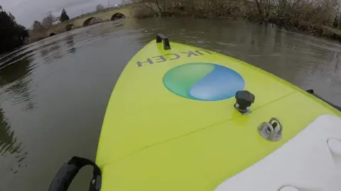 A view from the top of the boat, which shows it is in the middle of water. It is not far from a bridge and trees. 