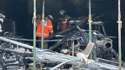 A close up showing the damage caused to a property after a fire. Three men wearing high vis orange jackets and helmets can be seen inside. There is lots of debris and a burnt up car in the shot.