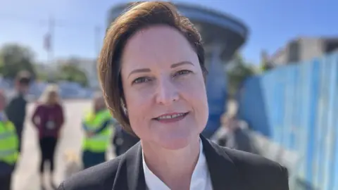 A woman with a brown geometric bob smiles at the camera. She is wearing a white silk shirt with a dark grey jacket 