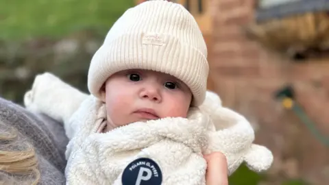 Baby Penny wearing a cream woollen hat and cream fleecy top looks towards camera. She is in her mother's arms outdoors.