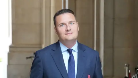 PA Media Wes Streeting looks directly at the camera as he walks outside. He is wearing a blue suit and blue tie, and a stone building is behind him. 
