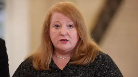 Naomi Long wearing a black top, with a silver necklace. She has long red hair