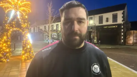 A man looking at the camera, he has dark hair and a beard. He is wearing a black jumper.