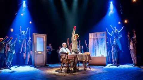 Tristram Kenton A middle-age man sits at a breakfast table. A young man in a white, ripped sweater stands next to him holding up a red envelope. They are surrounded by a cast of performers with their arms raised.