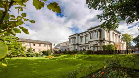 A large Victorian double-fronted white building with a 1990s style extended wing. The building sits on a sweeping lawn with a floral border.