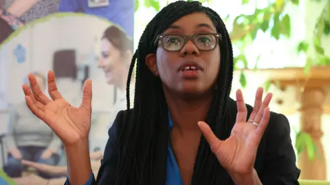 Kemi Badenoch holding her hands in the air. She is wearing a black blazer over a blue top, and has her mouth open while speaking in a sitting room at a hospice.