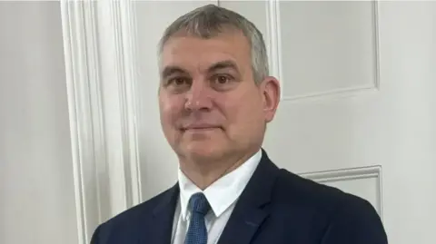 A man with short grey hair is looking into the camera and smiling. He is wearing a white shirt and blue tie and a blue jacket. He is standing in front of a white door.