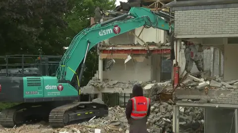 BBC A green bulldozer demolishing a white brick office block. A worker in a high visibility looks on. 