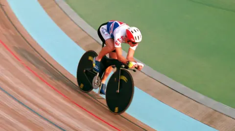 Action Images Film Chris Boardman on a bike racing around a velodrome circuit. He is wearing a red and white bike helmet and team GB's red, white, and blue colours on his top and bottom.