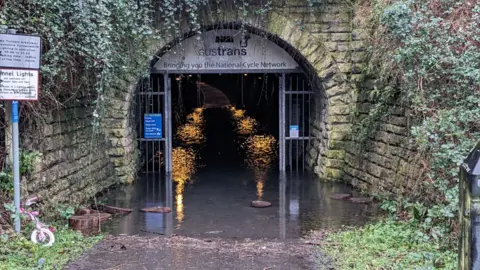 Two Tunnels Bath A closer shot of the metal gate and entrance to the Devonshire Tunnel. It is a long dark tunnel dimly lit by orange lights, which are reflecting on the floodwater that has pooled on the ground, stretching about 12 metres into the tunnel. 