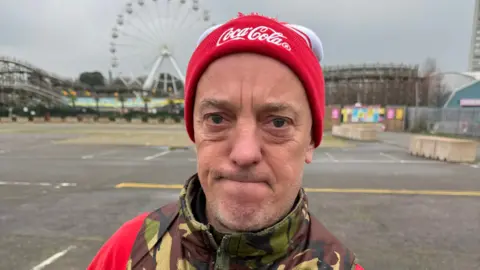 Keir Mackenzie/BBC A man in a red hat and a camouflage bodywarmer jacket. A rollercoaster and ferris wheel can be seen in the background.