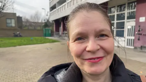 A middle-aged woman stands in front of a block of flats in Tower Hamlets and smiles into the camera