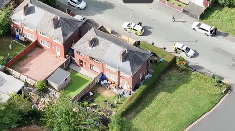 Greater Manchester Police Photograph of a house in north Manchester taken by a drone in the sky. The image shows police cars and officers searching the property.